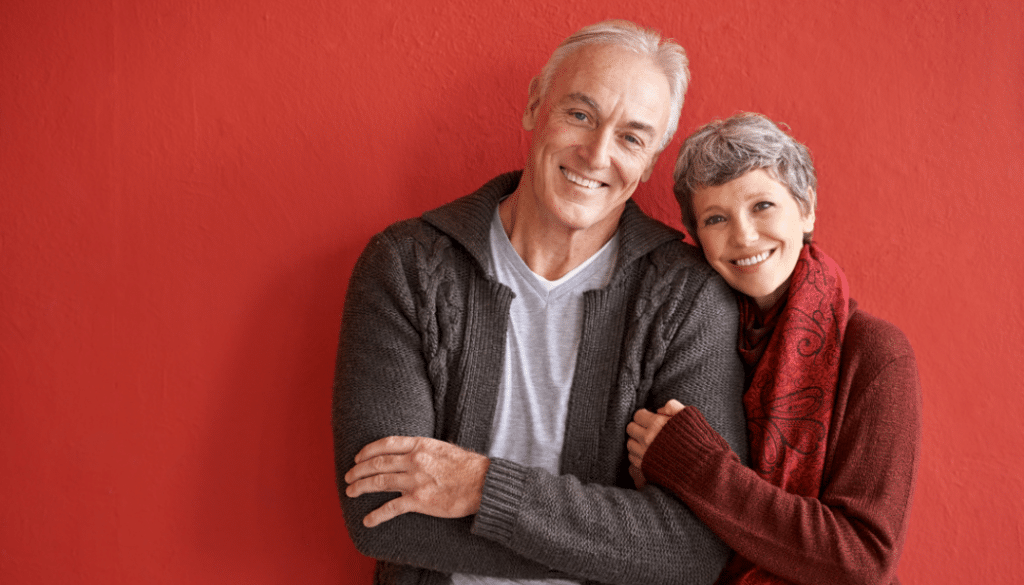 Older Male & Female embraced together looking into the camera with a red background. They are happy to signify happiness with aging.